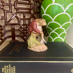 May include: A ceramic figurine of a frog wearing a red jacket and holding a shovel. The frog is sitting on a small mound of dirt with green grass. The figurine is sitting on a black book with gold lettering.