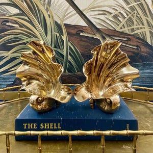 May include: Two gold-colored seashell sculptures, each with a textured, fan-like top and a rounded base. The sculptures are displayed on a blue book titled "THE SHELL". The background features a painting with tropical foliage.