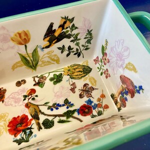 K&ouml;nnte beinhalten: Rechteckige Keramik-Backform mit wei&szlig;em Innenraum und hellgr&uuml;nem Rand und Griffen. Der Innenraum zeigt ein farbenfrohes Blumen- und Vogeldesign mit einer gelben Tulpe, roten Mohnblumen und einem gelb-schwarzen Vogel.
