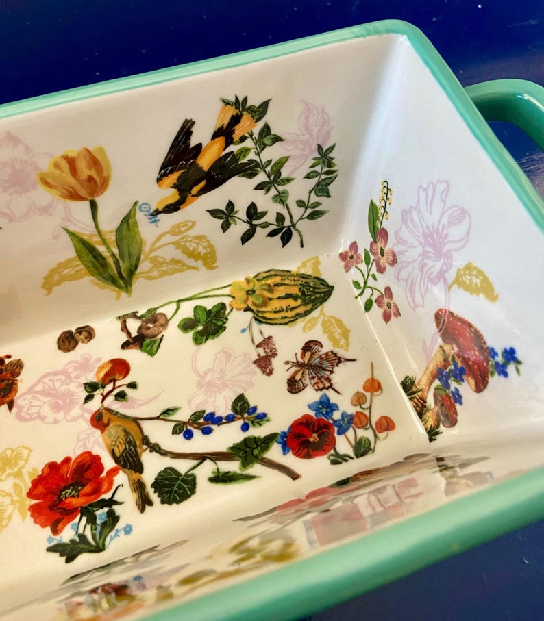 K&ouml;nnte beinhalten: Rechteckige Keramik-Backform mit wei&szlig;em Innenraum und hellgr&uuml;nem Rand und Griffen. Der Innenraum zeigt ein farbenfrohes Blumen- und Vogeldesign mit einer gelben Tulpe, roten Mohnblumen und einem gelb-schwarzen Vogel.