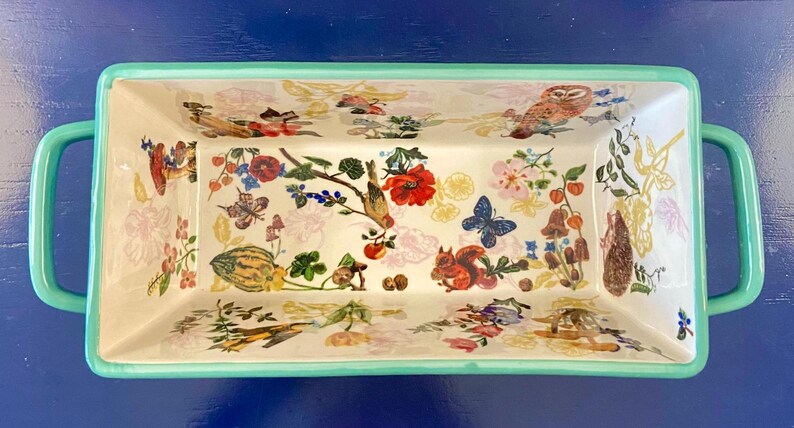 K&ouml;nnte beinhalten: Rechteckige Servierschale mit wei&szlig;em Innenraum und hellgr&uuml;nem Rand und Griffen. Der Innenraum ist mit farbenfrohen Illustrationen von Blumen, Schmetterlingen, V&ouml;geln und einem Eichh&ouml;rnchen verziert. Die Schale steht vor einem blauen Hintergrund.