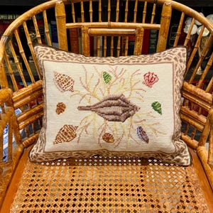 May include: A decorative pillow with a seashell design. The pillow features a cream-colored background with embroidered seashells, coral, and a central brown seashell. The border is a patterned brown fabric. The pillow is placed on a bamboo chair.