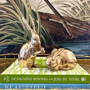 May include: Two polished brass rabbit figurines, one standing upright and one lying down, resting on a green book titled Designing Rooms with Joie de Vivre and a blue book titled Beautiful by Mark D. Sikes, featuring a metallic gold finish.