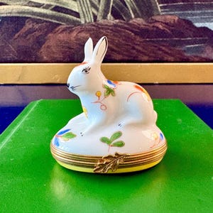 May include: A white porcelain trinket box shaped like a sitting rabbit. The rabbit features hand-painted floral motifs in green, orange, and blue. The base is secured with a gold-toned metal hinge and a decorative leaf-shaped clasp closure.