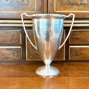May include: A large, polished silver-toned trophy cup featuring a smooth, tapered body, a wide circular base, and two tall, angular handles that extend from the rim down to the lower section of the vessel, creating a symmetrical silhouette.
