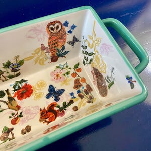 K&ouml;nnte beinhalten: Rechteckige Servierschale aus Keramik mit wei&szlig;em Innenraum und hellgr&uuml;nem Rand und Griffen. Die Schale ist mit farbenfrohen Illustrationen einer Eule, eines Eichh&ouml;rnchens, Schmetterlingen, Blumen und Pilzen verziert.