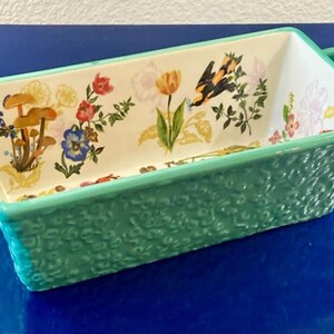 K&ouml;nnte beinhalten: Rechteckige Keramik-Backform mit floralem Design und t&uuml;rkisfarbener strukturierter Au&szlig;enseite. Der Innenraum zeigt bunte Blumen, Pilze und einen Schmetterling. Die Form hat zwei t&uuml;rkisfarbene Griffe.