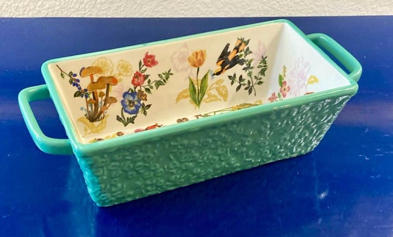 K&ouml;nnte beinhalten: Rechteckige Keramik-Backform mit floralem Design und t&uuml;rkisfarbener strukturierter Au&szlig;enseite. Der Innenraum zeigt bunte Blumen, Pilze und einen Schmetterling. Die Form hat zwei t&uuml;rkisfarbene Griffe.