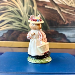 May include: A ceramic figurine of an anthropomorphic mouse wearing a cream-colored hat adorned with red berries and a blue accent. The mouse wears a white dress with red striped sleeves and carries a small, light-colored basket on its left arm.