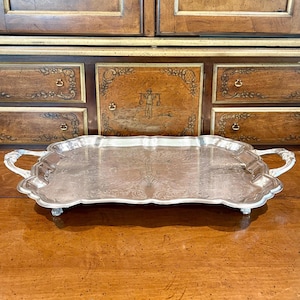 May include: A rectangular silver-toned serving tray with ornate, curved handles on each side. The tray features a scalloped edge and an intricate, embossed floral pattern across its flat surface, supported by four small, decorative feet.
