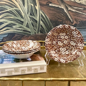 May include: A set of three ceramic bowls featuring a brown and white floral pattern. One bowl is displayed upright on a clear stand, while two bowls are stacked together. The bowls have a white interior and a detailed brown floral exterior.
