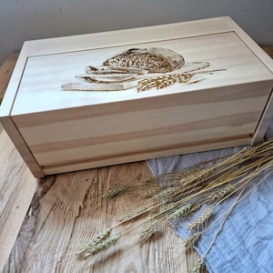 Puede incluir: Una caja de pan de madera con una tapa deslizante. La tapa tiene un diseño de pan y espigas de trigo. La caja está sobre una superficie de madera con un paño azul y espigas de trigo.