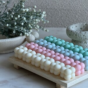 May include: A selection of pastel-coloured bubble candles in white, pink, blue, and green, displayed on a light wooden tray. The candles have a distinctive spherical shape. Decorative items, including a vase and flowers, are in the background.