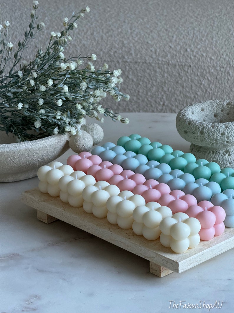 May include: A selection of pastel-coloured bubble candles in white, pink, blue, and green, displayed on a light wooden tray. The candles have a distinctive spherical shape. Decorative items, including a vase and flowers, are in the background.
