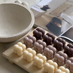 May include: A selection of bubble-shaped candles in cream, mauve, and brown hues, displayed on a rectangular wooden tray. An off-white textured bowl and a magazine are in the background. The candles are unscented and decorative.