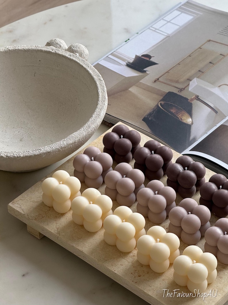 May include: A selection of bubble-shaped candles in cream, mauve, and brown hues, displayed on a rectangular wooden tray. An off-white textured bowl and a magazine are in the background. The candles are unscented and decorative.
