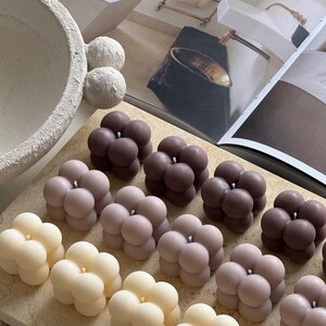 May include: A selection of bubble-shaped candles in cream, mauve, and brown hues. The candles are displayed on a light-toned surface, with a magazine and decorative bowl in the background. The candles have a modern, minimalist design.