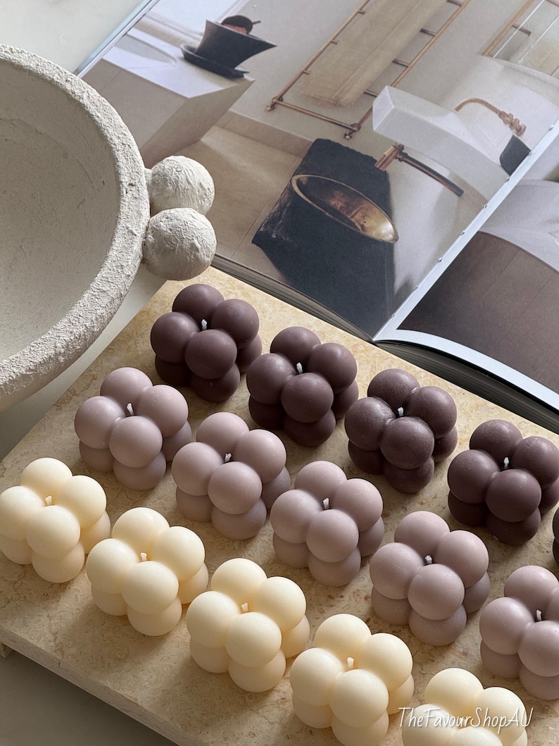 May include: A selection of bubble-shaped candles in cream, mauve, and brown hues. The candles are displayed on a light-toned surface, with a magazine and decorative bowl in the background. The candles have a modern, minimalist design.
