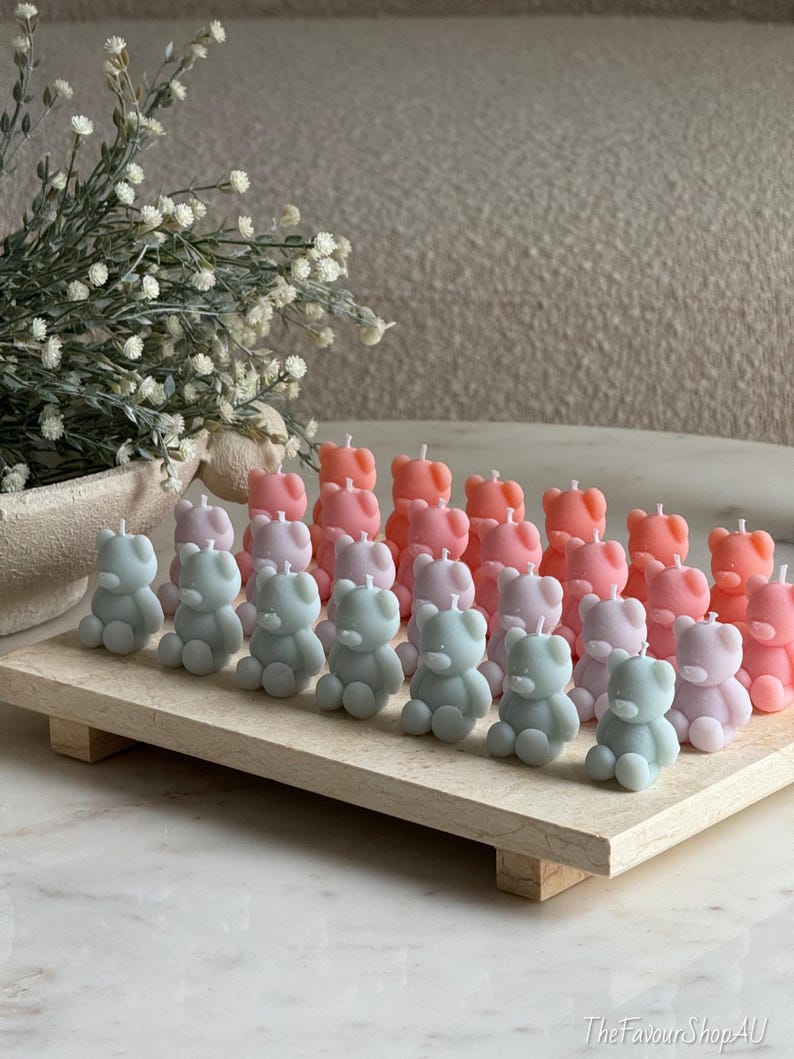 May include: A collection of small, bear-shaped candles in pastel shades of pink, lavender, and sage green, arranged in rows on a rectangular stone tray. Each candle features a single white wick at the top. The text TheFavourShopAU is visible.