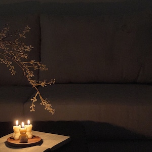 May include: Three small, cream-colored bear-shaped candles are lit and placed on a circular wooden tray. The tray rests on an open book with visible text. A delicate, thin branch with small buds extends over the candles in a dimly lit setting.