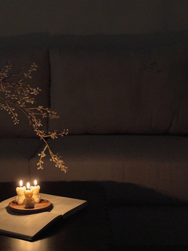 May include: Three small, cream-colored bear-shaped candles are lit and placed on a circular wooden tray. The tray rests on an open book with visible text. A delicate, thin branch with small buds extends over the candles in a dimly lit setting.