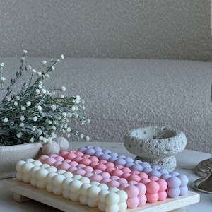 May include: A selection of bubble candles in a gradient of colours, from white to pink and purple, arranged on a light wooden tray. The candles have a spherical design. The background includes a neutral-toned sofa and decorative elements.