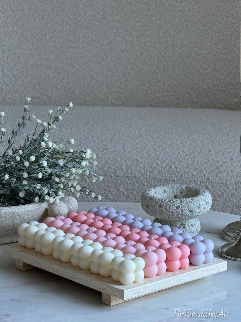 May include: A selection of bubble candles in a gradient of colours, from white to pink and purple, arranged on a light wooden tray. The candles have a spherical design. The background includes a neutral-toned sofa and decorative elements.