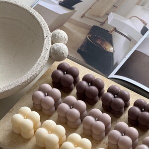 May include: A selection of bubble-shaped candles in cream, pale pink, and dark brown. The candles are arranged on a light-coloured rectangular surface. The background includes a decorative bowl, spheres, and a magazine.
