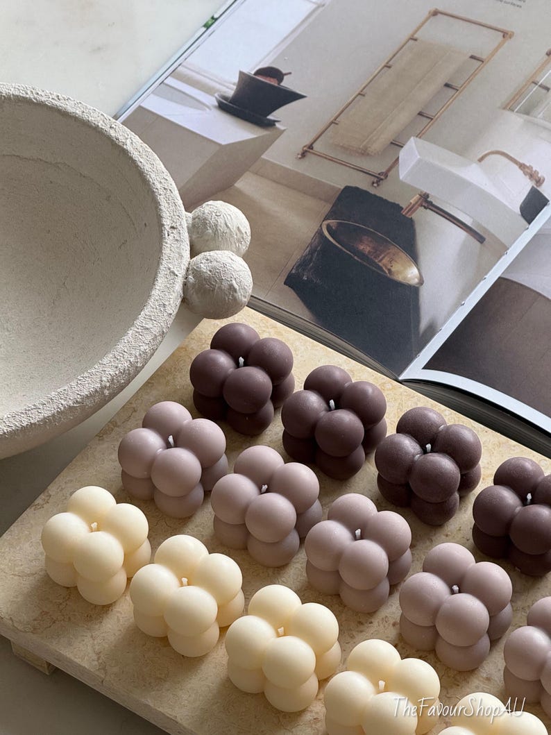 May include: A selection of bubble-shaped candles in cream, pale pink, and dark brown. The candles are arranged on a light-coloured rectangular surface. The background includes a decorative bowl, spheres, and a magazine.