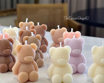 Peut inclure: Une collection de petites bougies en forme d'ours dans diverses couleurs pastel et neutres, notamment blanc, crème, beige, marron et violet clair. Chaque bougie possède une mèche blanche au sommet. Le texte TheFavourShopAU est visible.
