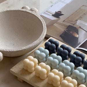 May include: A selection of bubble candles in cream, light blue, and dark blue hues, displayed on a rectangular wooden tray. A textured, light-coloured bowl and a magazine are in the background. These candles are a decorative home accessory.