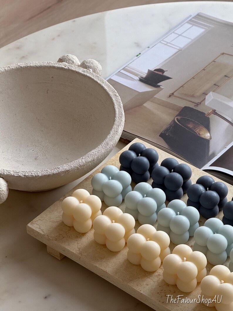 May include: A selection of bubble candles in cream, light blue, and dark blue hues, displayed on a rectangular wooden tray. A textured, light-coloured bowl and a magazine are in the background. These candles are a decorative home accessory.