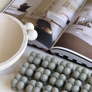 May include: Several grey bubble candles, each composed of clustered spheres, are arranged in rows. A decorative bowl and white spheres are in the background, along with an open magazine. The Favour Shop AU is printed on the bottom right.