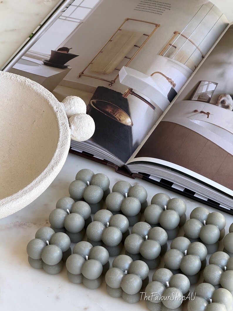 May include: Several grey bubble candles, each composed of clustered spheres, are arranged in rows. A decorative bowl and white spheres are in the background, along with an open magazine. The Favour Shop AU is printed on the bottom right.