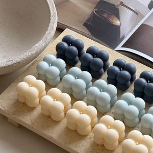 May include: A selection of bubble-shaped candles in cream, light blue, and dark grey, displayed on a rectangular, light-coloured tray. Each candle features a single wick. The background includes a decorative bowl and a magazine.