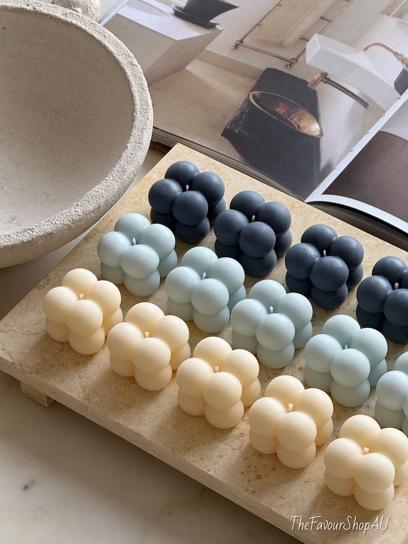 May include: A selection of bubble-shaped candles in cream, light blue, and dark grey, displayed on a rectangular, light-coloured tray. Each candle features a single wick. The background includes a decorative bowl and a magazine.