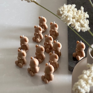 May include: A collection of eleven small, light brown, bear-shaped candles, each featuring a single white wick at the top of the head. The bears are depicted in a seated position with rounded ears and limbs, arranged in a cluster on a smooth surface.