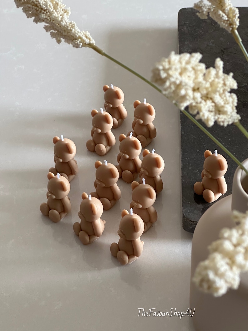 May include: A collection of eleven small, light brown, bear-shaped candles, each featuring a single white wick at the top of the head. The bears are depicted in a seated position with rounded ears and limbs, arranged in a cluster on a smooth surface.