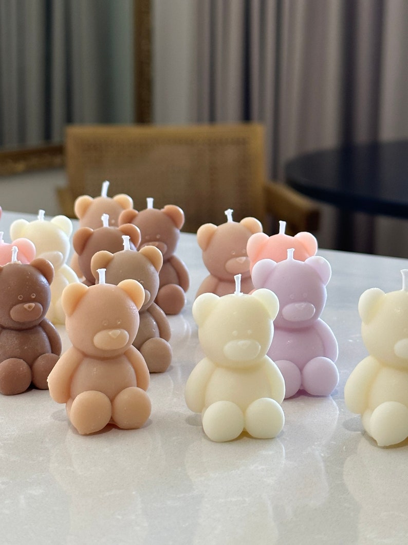 May include: A collection of small, molded bear-shaped candles in various pastel and neutral colors including cream, light brown, tan, and soft lavender. Each candle features a simple, smooth design with a single white wick centered on the head.
