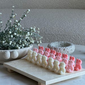 May include: A collection of small, bear-shaped candles in shades of cream and soft pink, arranged in rows on a rectangular stone tray. The candles feature a simple wick at the top. The text TheFavourShopAU is visible in the bottom right corner.