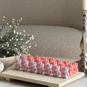 May include: A collection of small, bear-shaped candles in shades of pastel pink, peach, and lavender, arranged in rows on a rectangular stone tray. Each candle features a single white wick at the top of the head, with a smooth, matte finish.