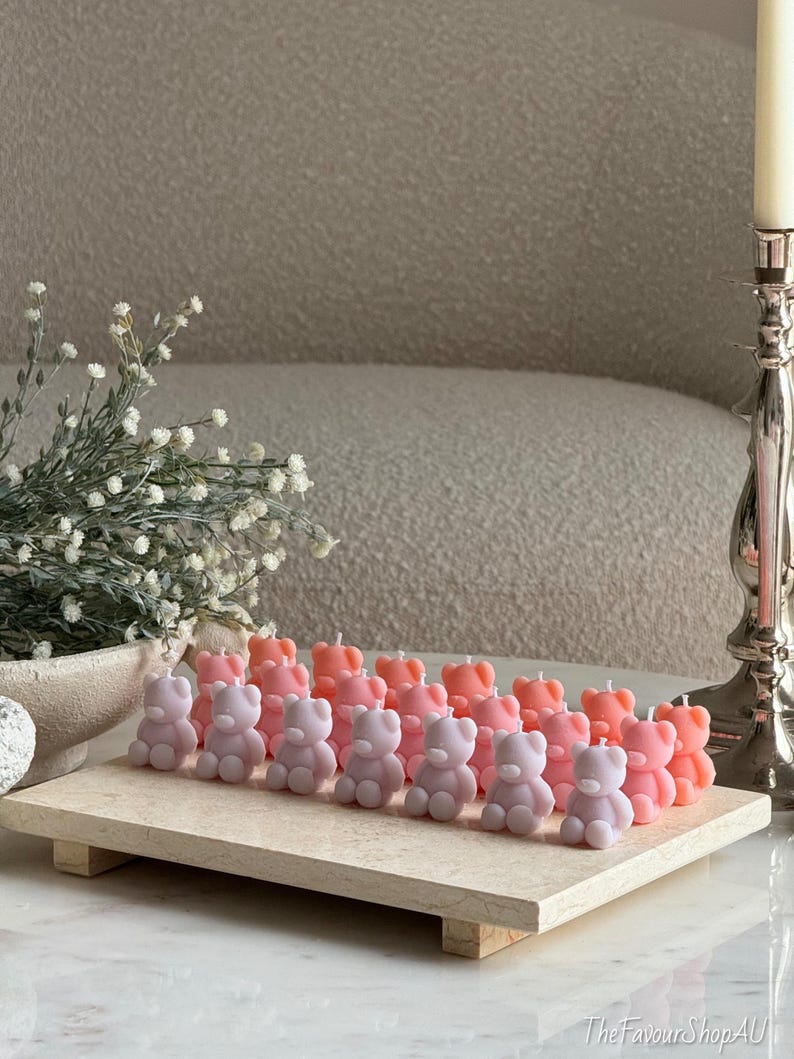 May include: A collection of small, bear-shaped candles in shades of pastel pink, peach, and lavender, arranged in rows on a rectangular stone tray. Each candle features a single white wick at the top of the head, with a smooth, matte finish.