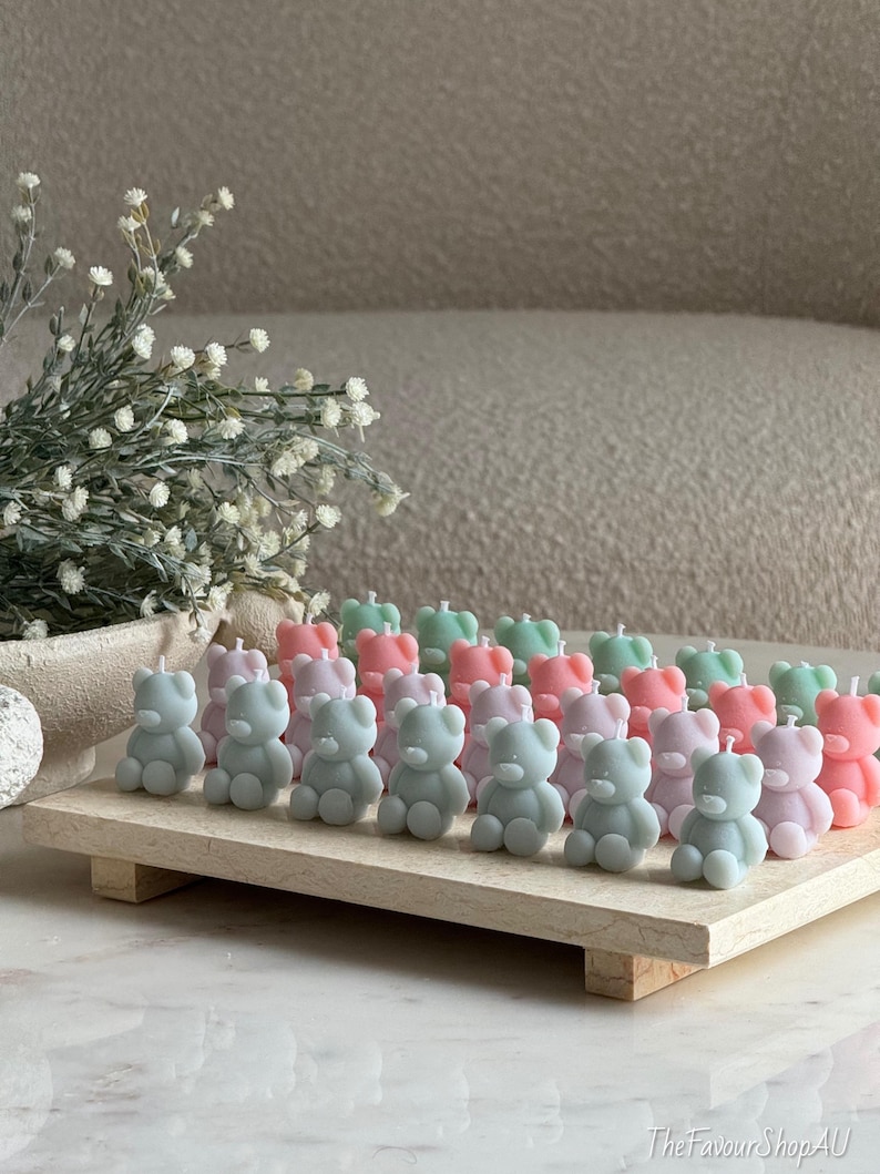 May include: A collection of small, bear-shaped candles in pastel pink, light blue, and sage green arranged in rows on a rectangular stone tray. Each candle features a single white wick at the top. The text TheFavourShopAU is visible at the bottom.