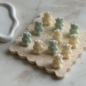 May include: A collection of ten small bear-shaped candles in alternating cream and pale green colors, each featuring a single centered wick. The candles are arranged on a square, porous, textured beige base with scalloped edges. Text reads TheFavourShopAU.