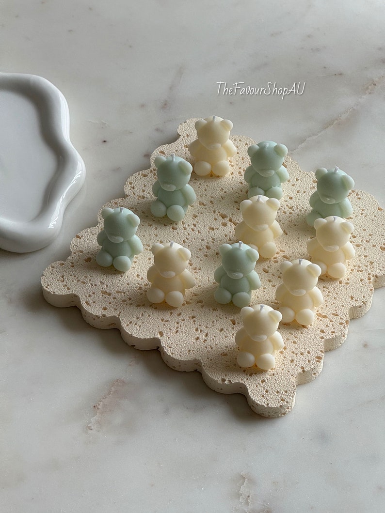 May include: A collection of ten small bear-shaped candles in alternating cream and pale green colors, each featuring a single centered wick. The candles are arranged on a square, porous, textured beige base with scalloped edges. Text reads TheFavourShopAU.