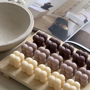 May include: A selection of bubble-shaped candles in cream, beige, and brown hues, displayed on a light wooden tray. The candles feature a smooth, rounded design. The background includes a decorative bowl and a magazine.