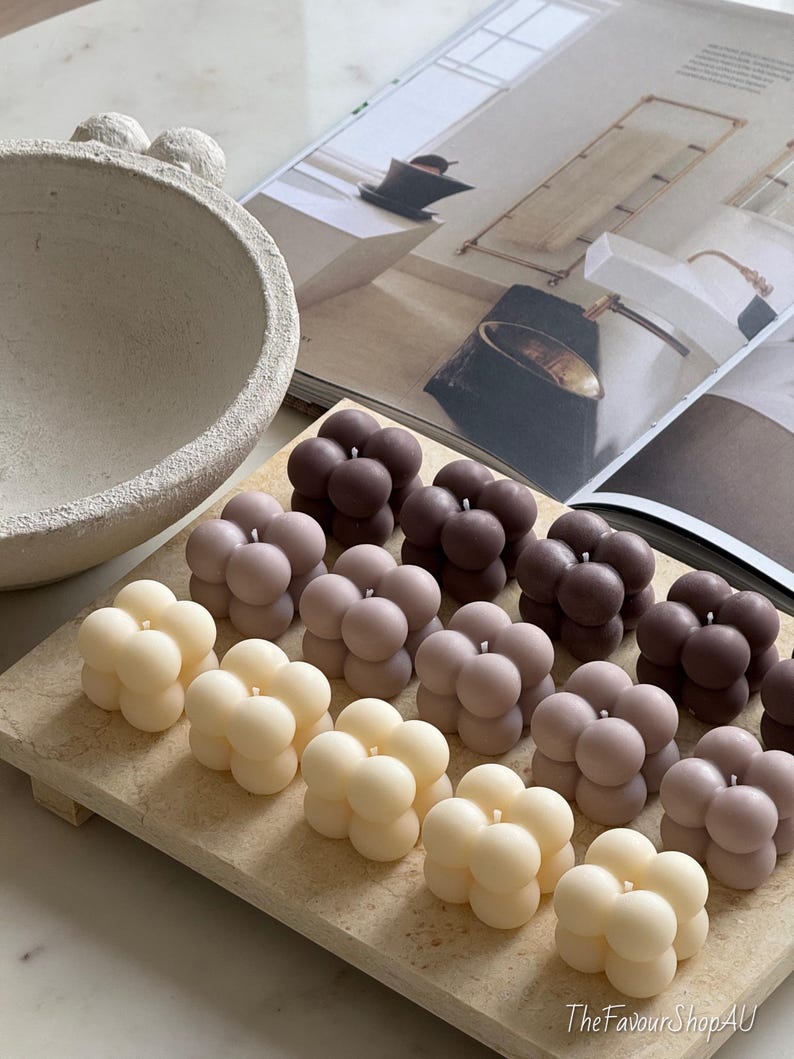 May include: A selection of bubble-shaped candles in cream, beige, and brown hues, displayed on a light wooden tray. The candles feature a smooth, rounded design. The background includes a decorative bowl and a magazine.