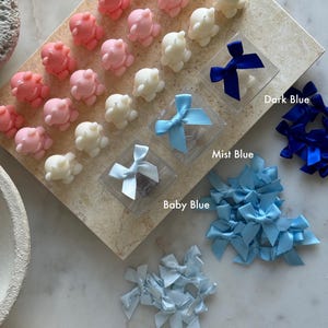 May include: A selection of octopus-shaped candles in pink and white, presented on a rectangular surface. Clear boxes hold blue satin bows in dark, mist, and baby blue. Loose bows in similar shades are also visible.