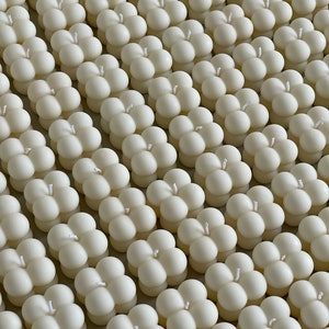 May include: A top-down view of many ivory bubble candles, neatly aligned. Each candle features four spheres and a central white wick. The candles have a smooth, matte surface, giving a consistent, soft look. The arrangement suggests a collection of decorative items.