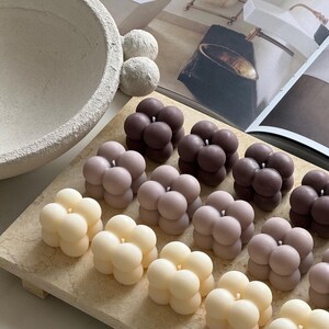 May include: A selection of bubble-shaped candles in cream, mauve, and brown hues. The candles are displayed on a light-toned rectangular tray. A textured bowl and decorative spheres are visible in the background.